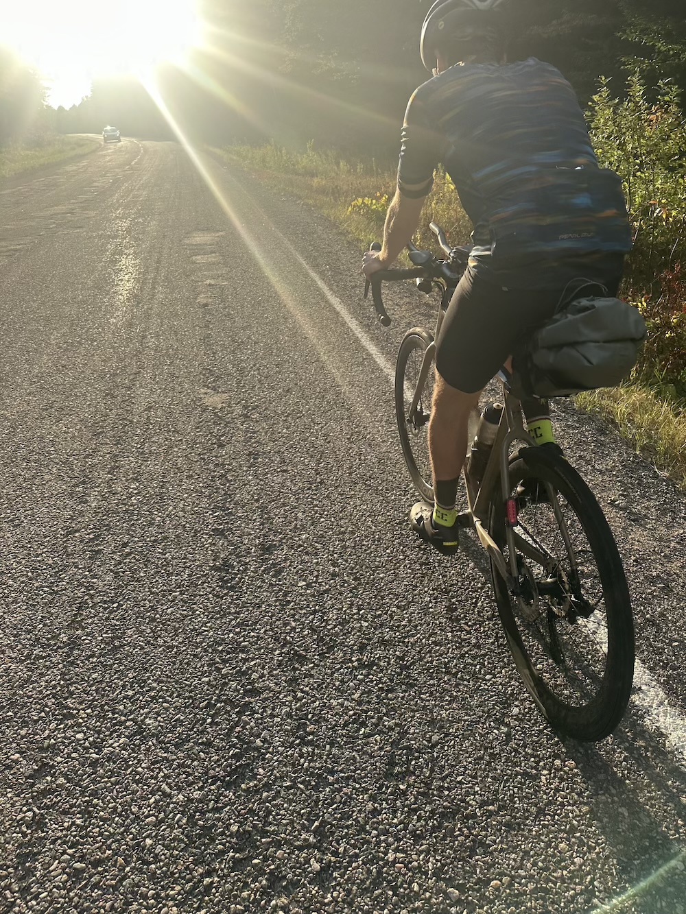 A cyclist riding uphill into the sun on a chipseal road surrounded by pine forests.