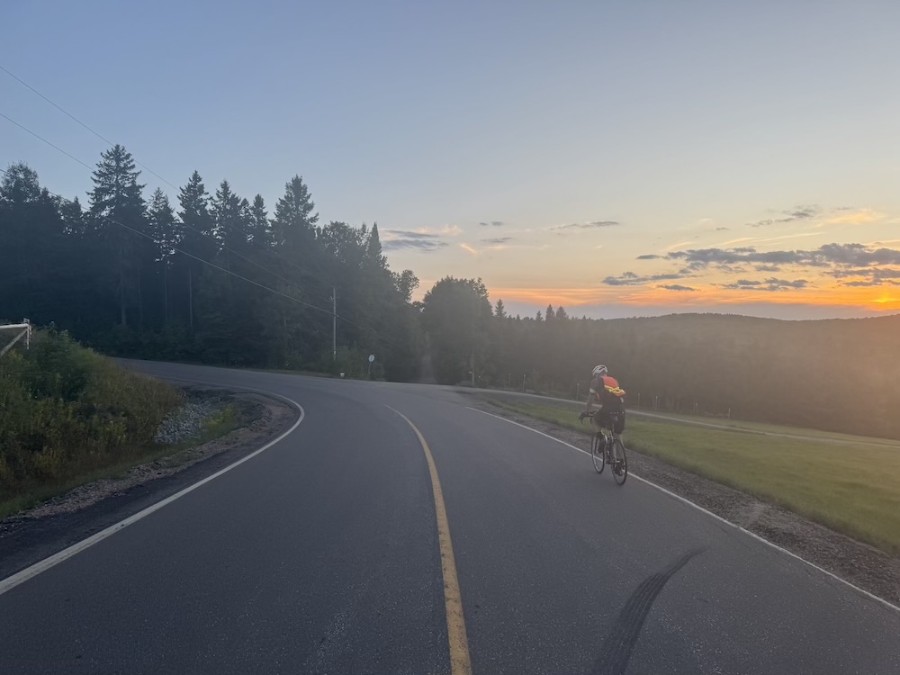 A cyclist riding in the country at sunset approaching a curvy uphill section.