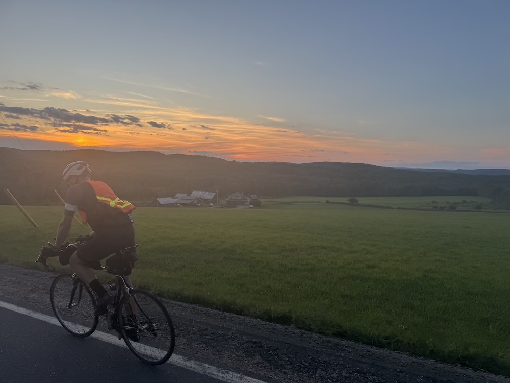 A cyclist rides along the ridge of a farm road at sunset, with a farm in view below.