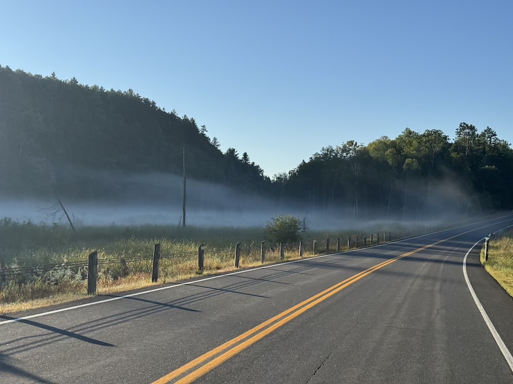 Fog lays amongst the trees in a valley near Bancroft, Ontario, with an uphill road in the distance.