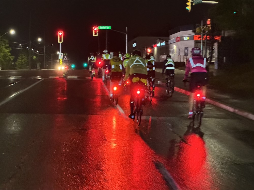 A group of 30 or more cyclists wearing reflective gear and with red tail lights riding on wet roads in Barrie, Ontario
