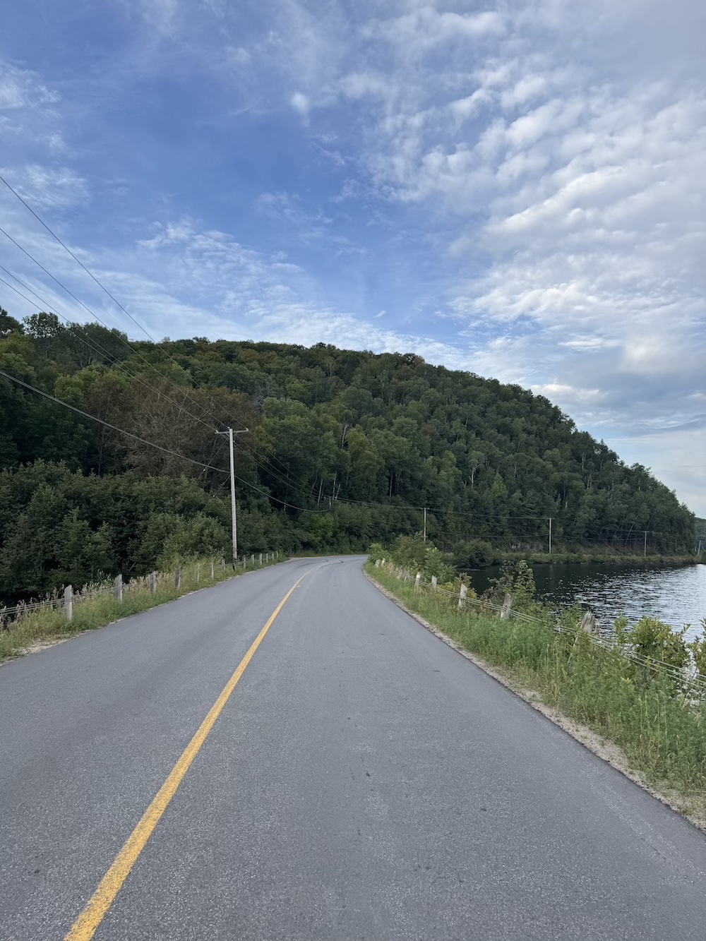 A road along a lake with a pine-covered hill in the background