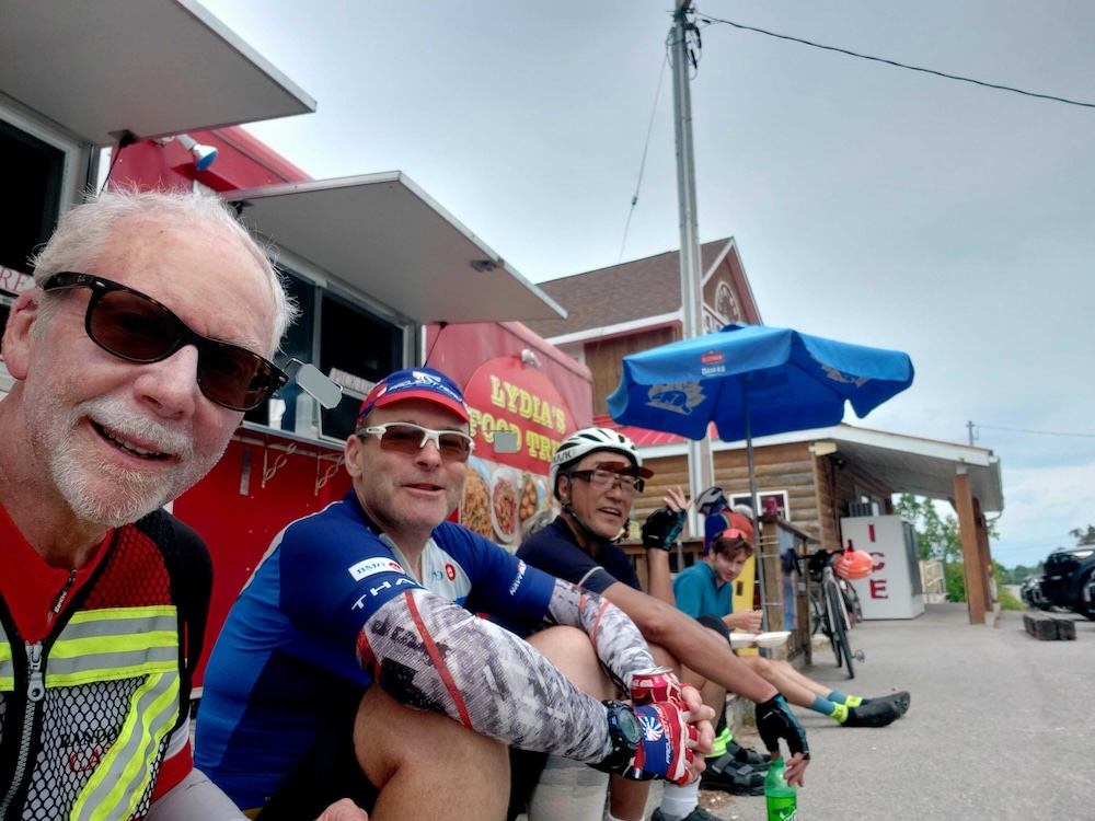 A group of cyclists sitting on the edge of a deck eating takeout food.