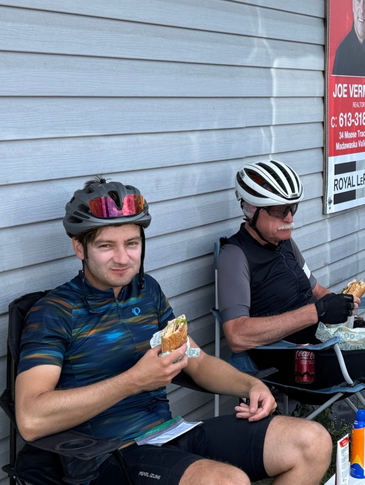 Two cyclists sitting in camping chairs in the shade of a building eating subway sandwiches in their cycling kit.