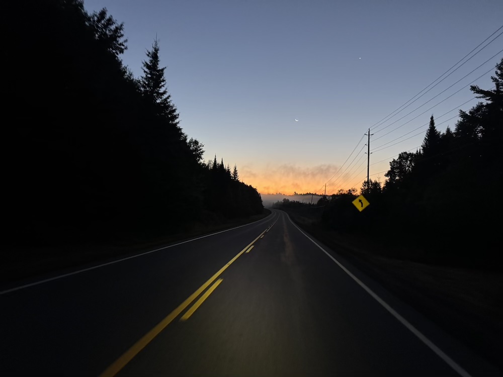 The sun rising amongst pine trees and Canadian shield at the end of a road with more uphill in the horizon.