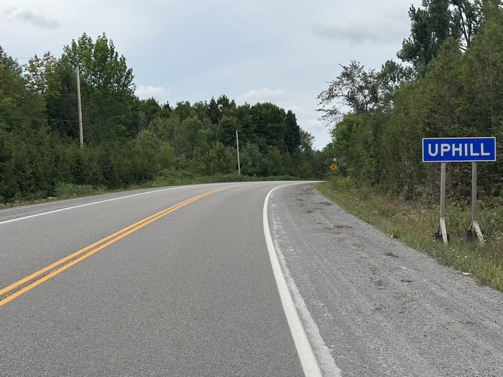 A road approaching the town sign for a town called Uphill, Ontario.